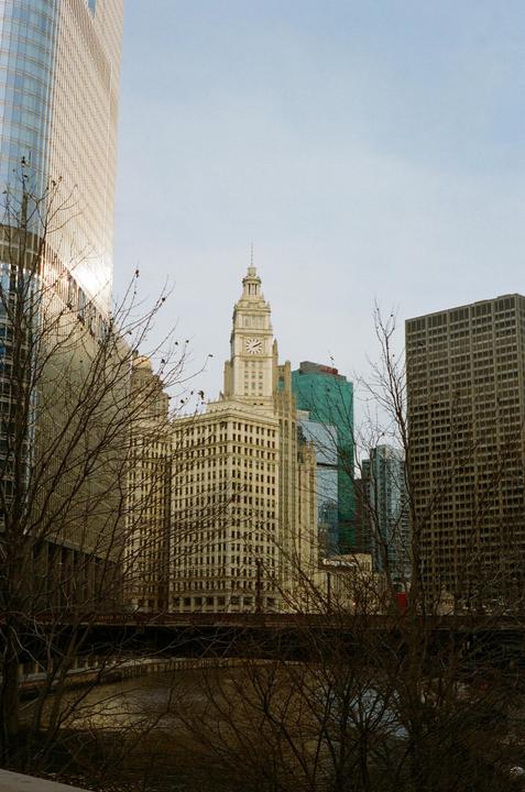 A clock tower rises above other buildings and bare trees next to a river.