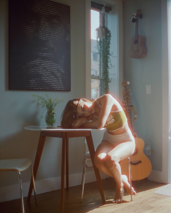 A woman rests her head on a table in a sunlit room with a guitar and ukulele.