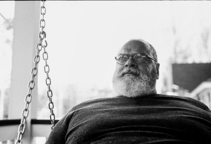 A man with a beard and glasses sits on a porch swing, looking up and to the right.