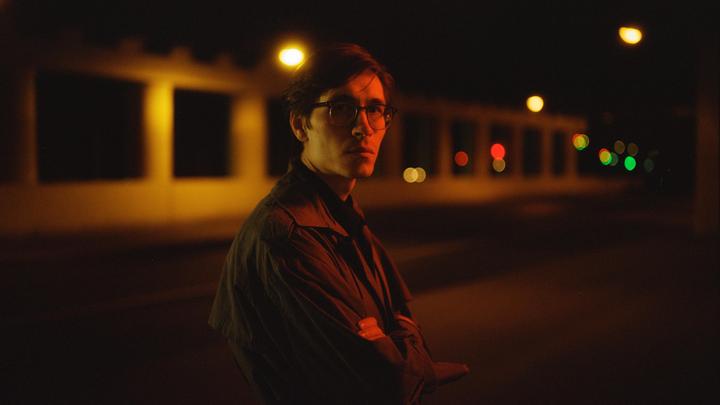 A man wearing glasses and a coat stands with his arms crossed at night, illuminated by streetlights.