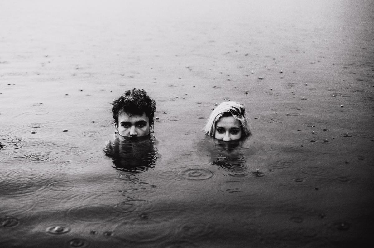 A man and a woman are submerged in water with only their heads visible, as rain falls around them.