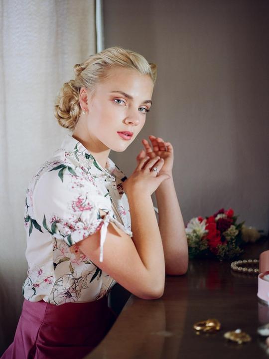 A woman with braided hair and a floral shirt sits at a wooden table with her hands clasped.