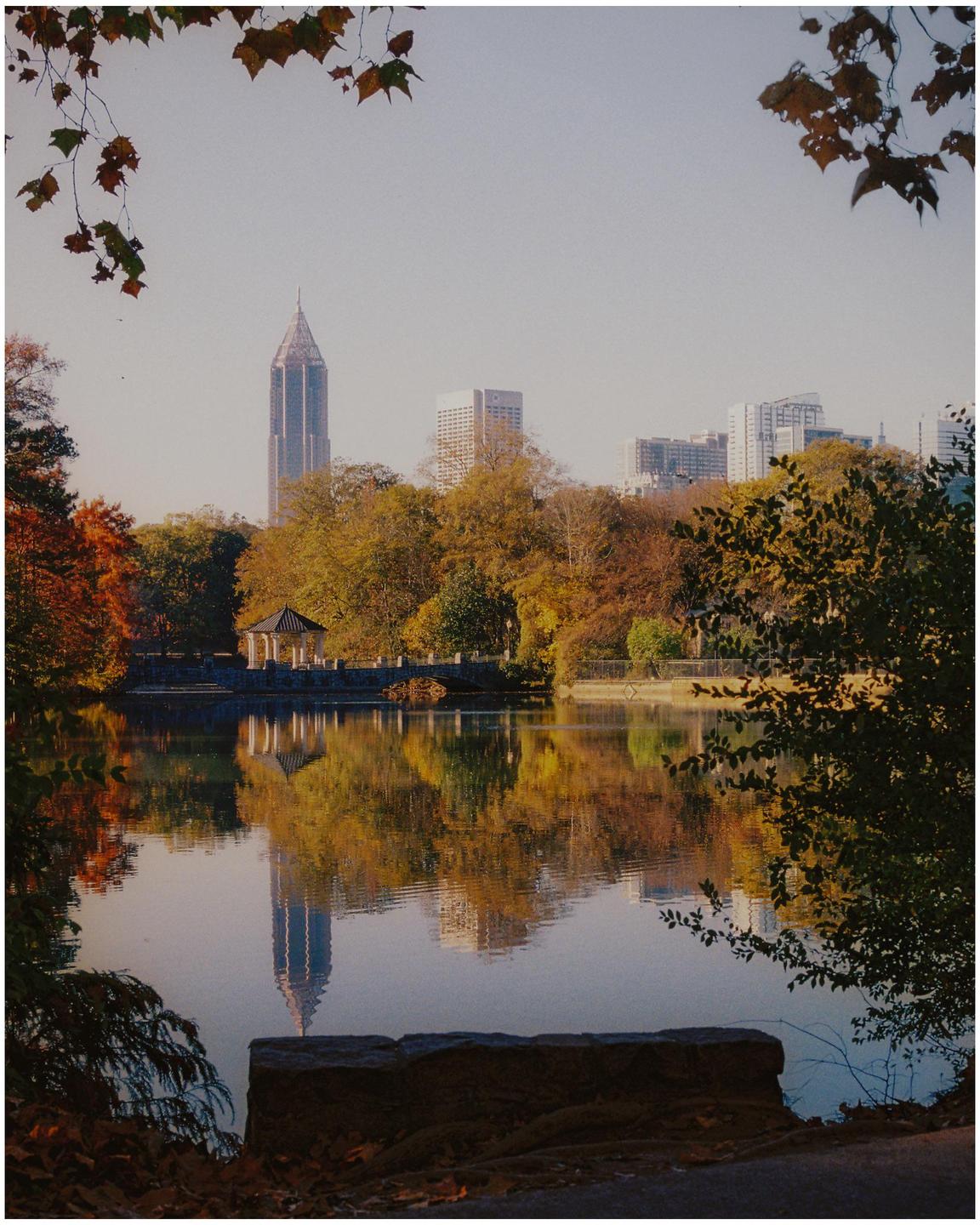 A city skyline with a prominent skyscraper is reflected in a calm lake surrounded by autumn trees and a gazebo.