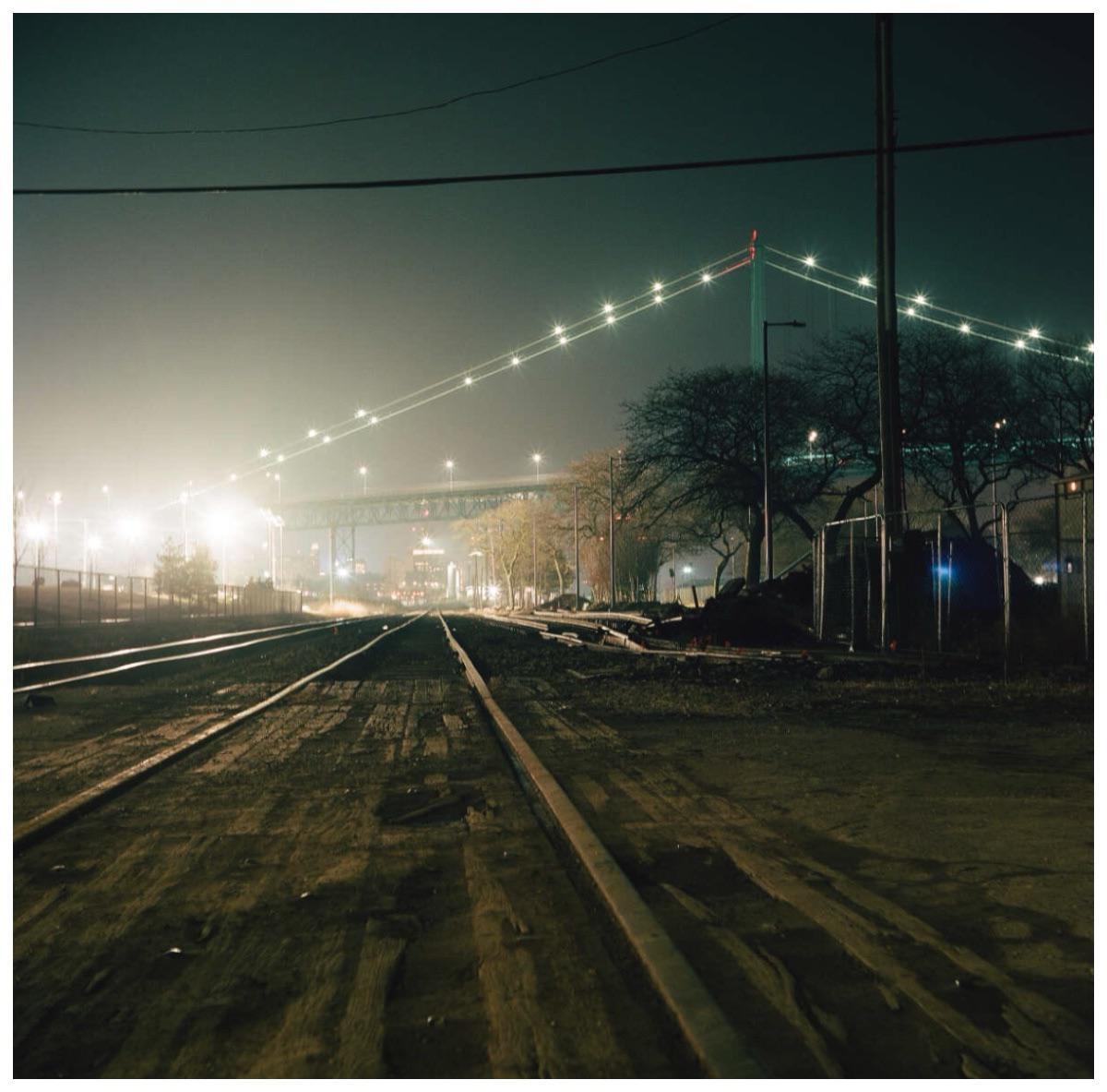 Railroad tracks lead towards a brightly lit bridge in the distance at night.