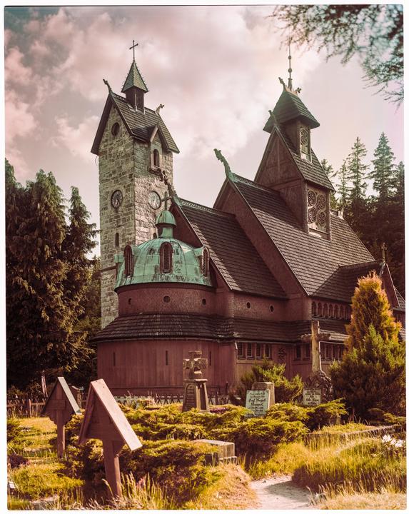 A wooden church with a stone tower and green domes stands among trees and gravestones under a cloudy sky.