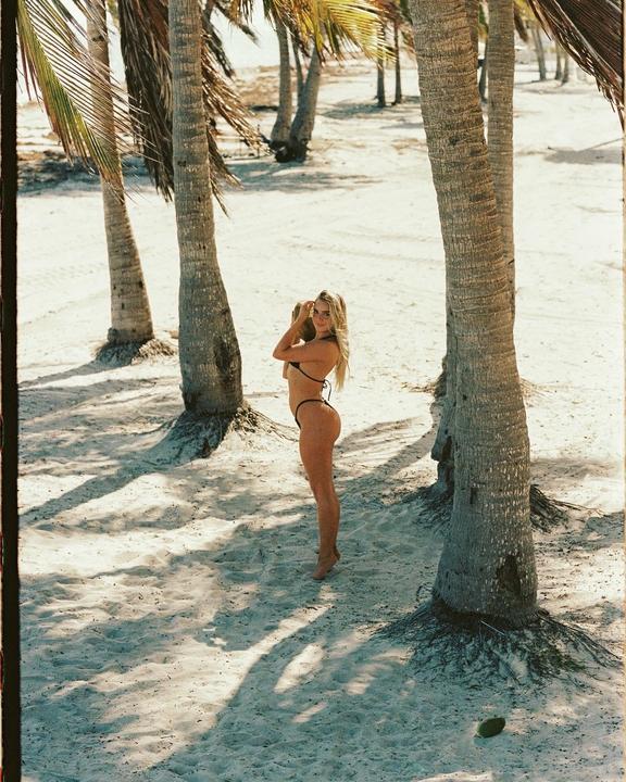 A woman in a bikini stands on a sandy beach among palm trees, holding a coconut.