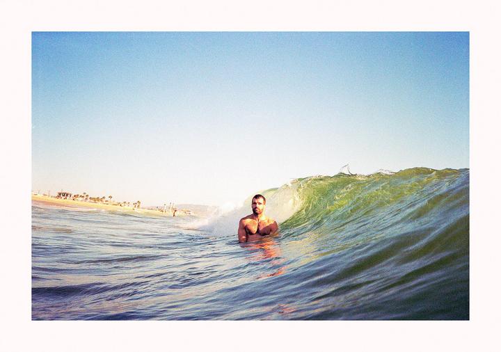 A man is in the ocean with a wave breaking behind him and a beach in the distance.