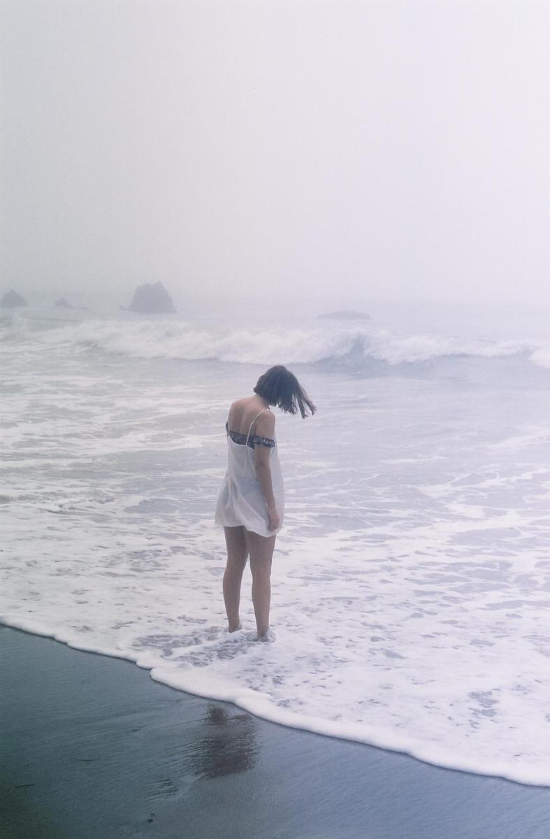 A woman stands in the foamy surf of a hazy beach with rocks in the distance.