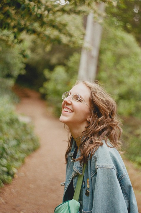 A young woman with glasses and a denim jacket smiles while looking up, standing on a dirt path surrounded by green foliage.