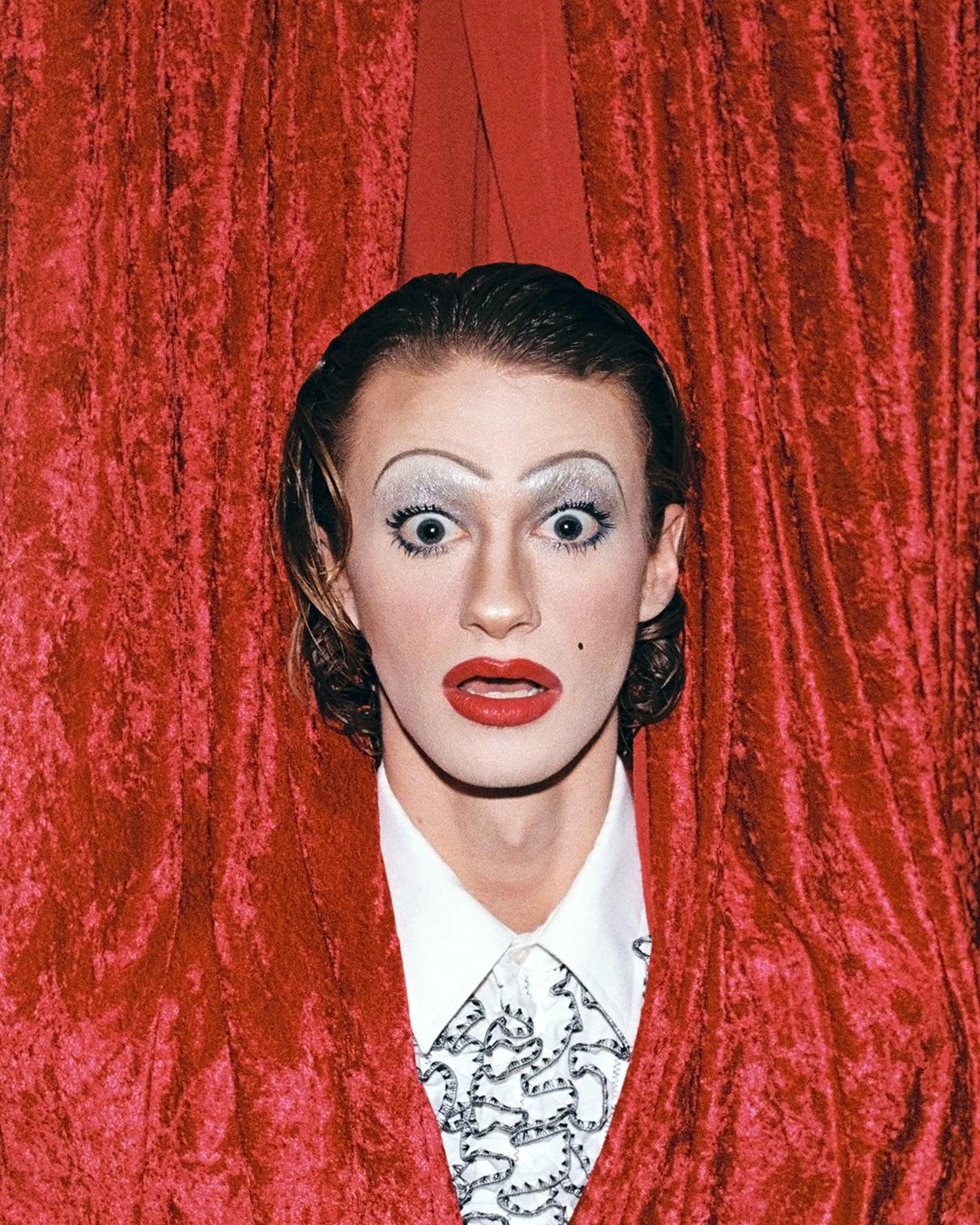 A person with dramatic makeup and a red velvet curtain looks directly at the viewer.