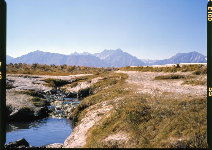A small stream flows through a grassy, rocky landscape with mountains in the distance under a clear sky.