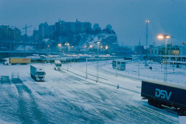 Several trucks and a bus drive on a snow-covered road with buildings and a hill in the background.