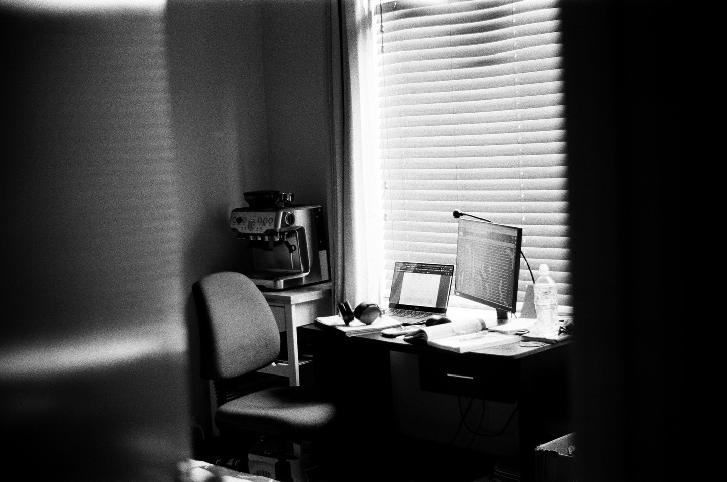 Sunlight streams through blinds onto a desk with a laptop, monitor, and headphones, next to a coffee machine and chair.