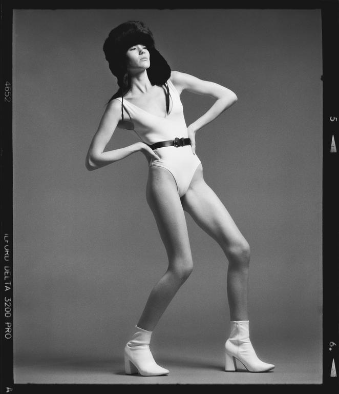 A woman in a white bodysuit, black belt, white boots, and a fur hat poses with her hands on her hips.