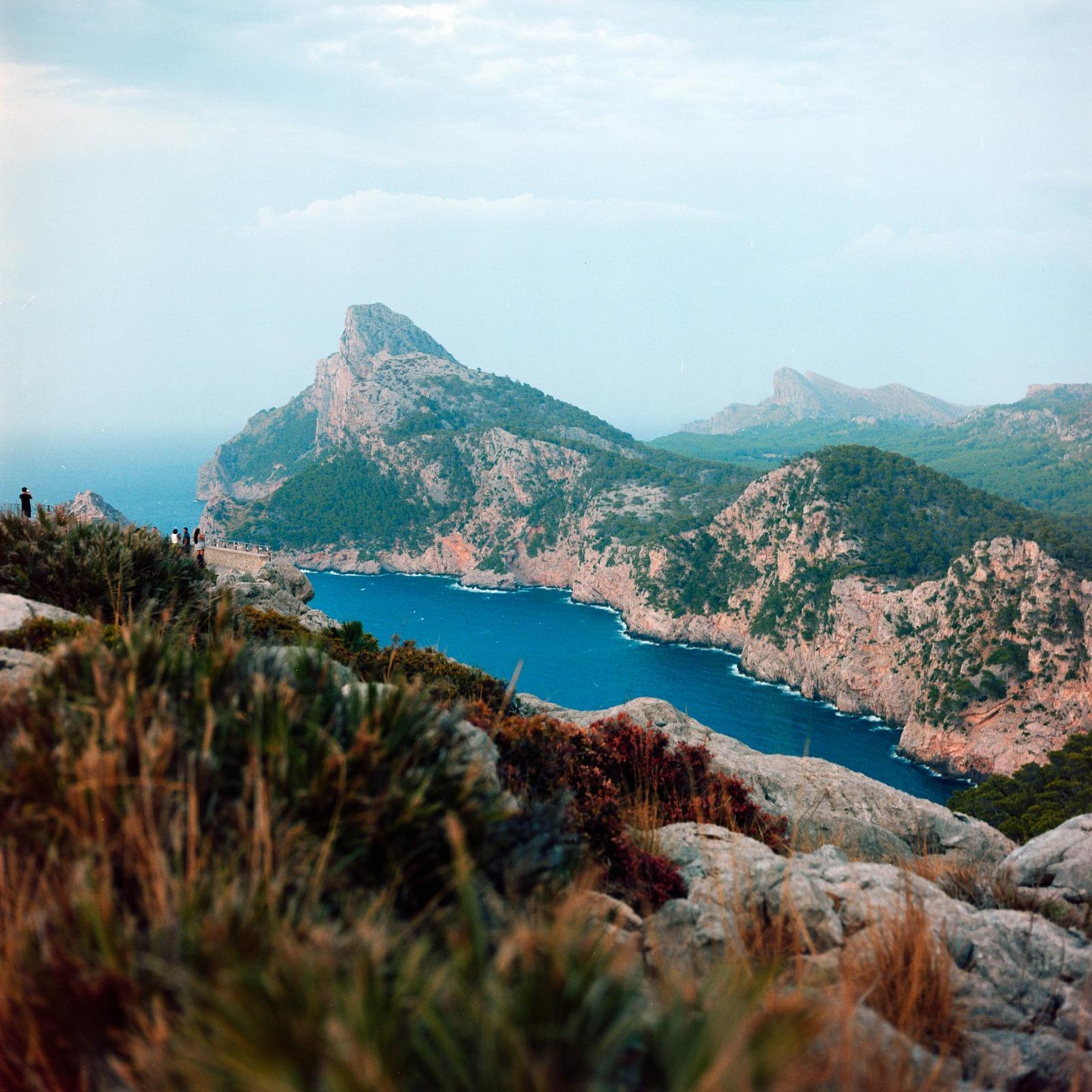 A winding bay with blue water is surrounded by rocky cliffs and green trees under a cloudy sky.