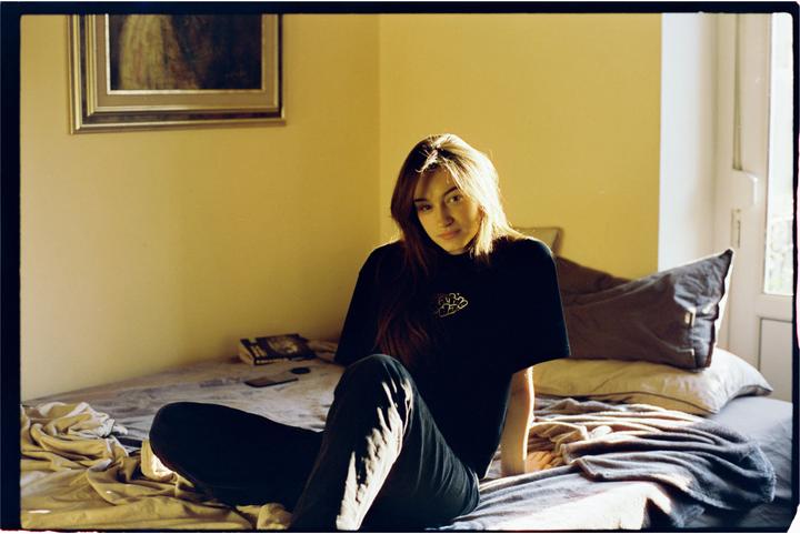 A woman sits on a bed in a sunlit room, looking towards the viewer.