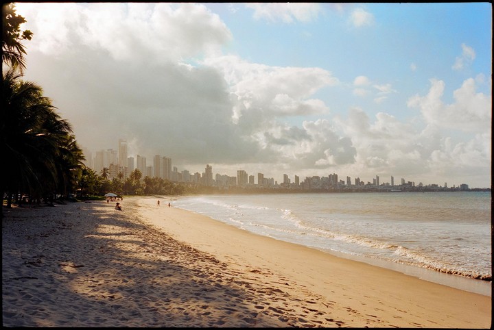 A wide sandy beach with palm trees on the left and a city skyline in the distance under a cloudy sky.