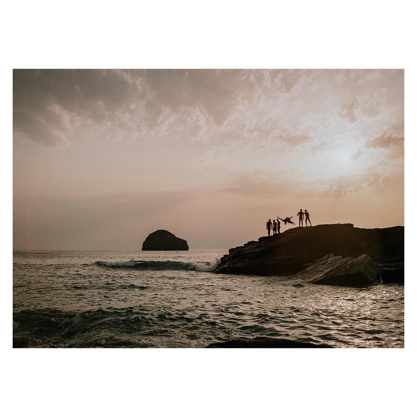 A group of people stand on a rocky outcrop by the ocean at sunset, with one person jumping in the air.