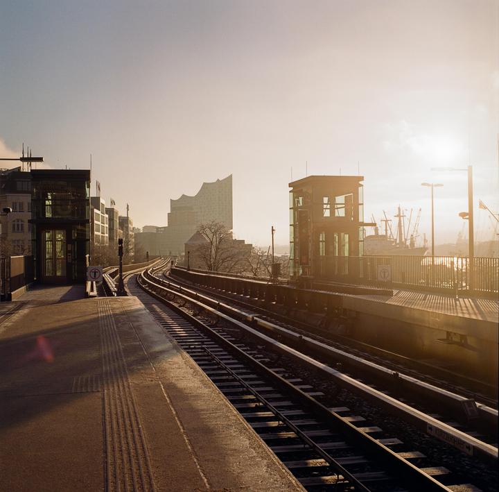A train station platform and tracks curve towards a city skyline with a prominent modern building and a ship in the distance under a hazy sky.