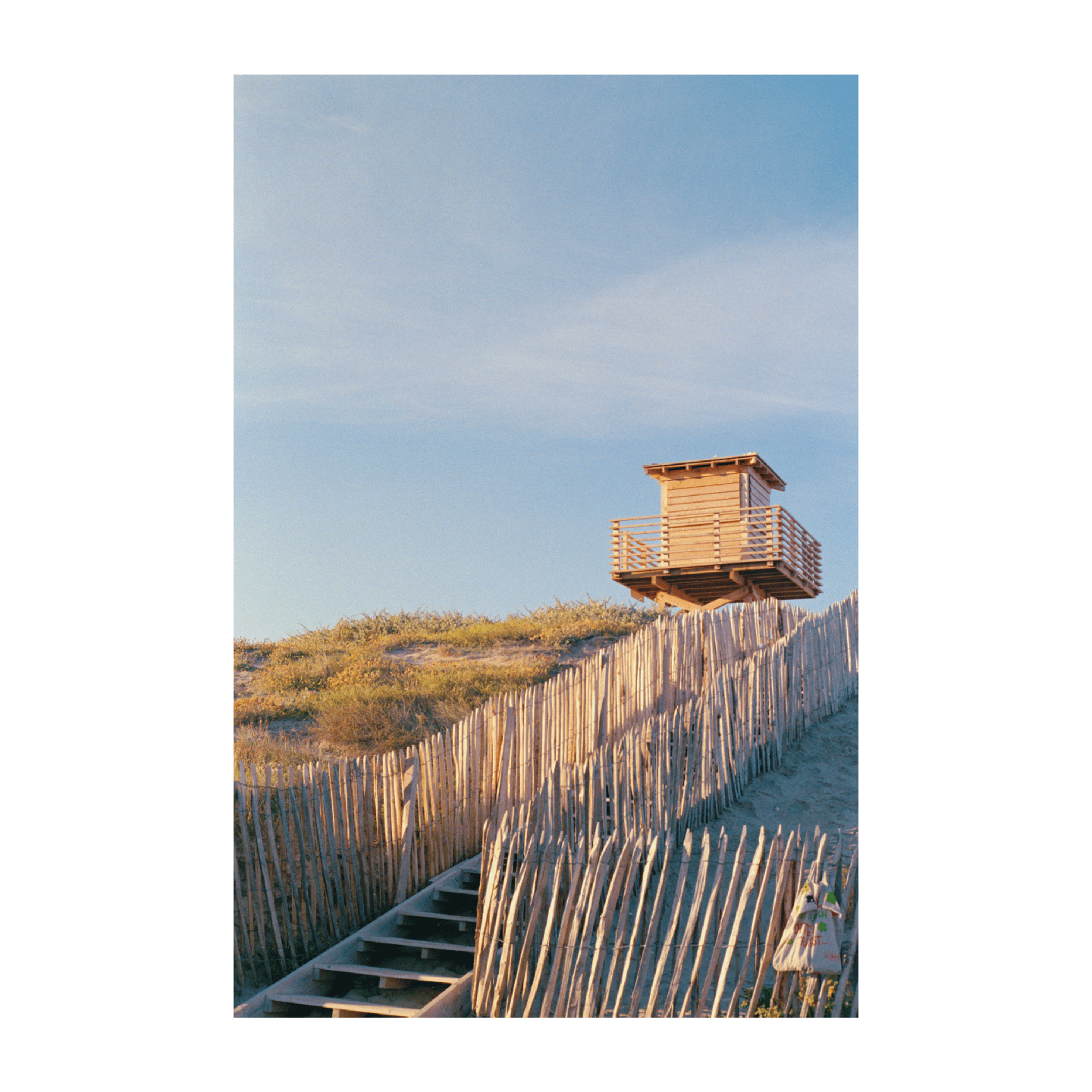 A wooden lifeguard tower stands on a sandy dune with a wooden fence and stairs leading up to it.