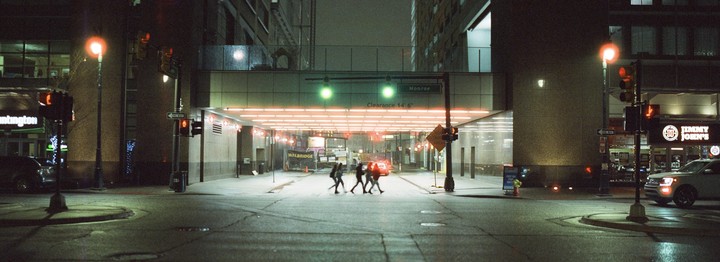 Four people cross a city street at night, illuminated by streetlights and neon signs.