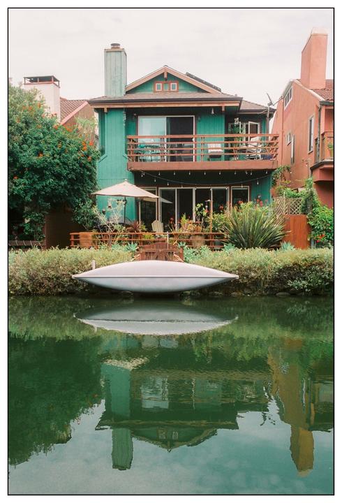 A teal house with a balcony and patio overlooks a canal with a small boat and its reflection.