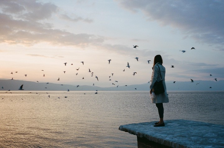 A woman stands on a concrete pier looking out at a lake with many birds flying over the water at dusk.