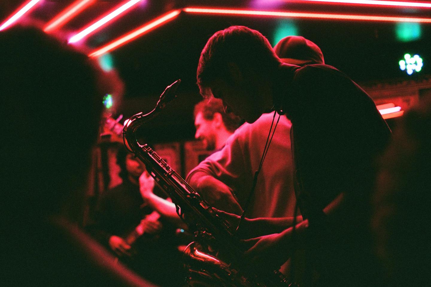A man plays a saxophone on a stage lit by red neon lights.