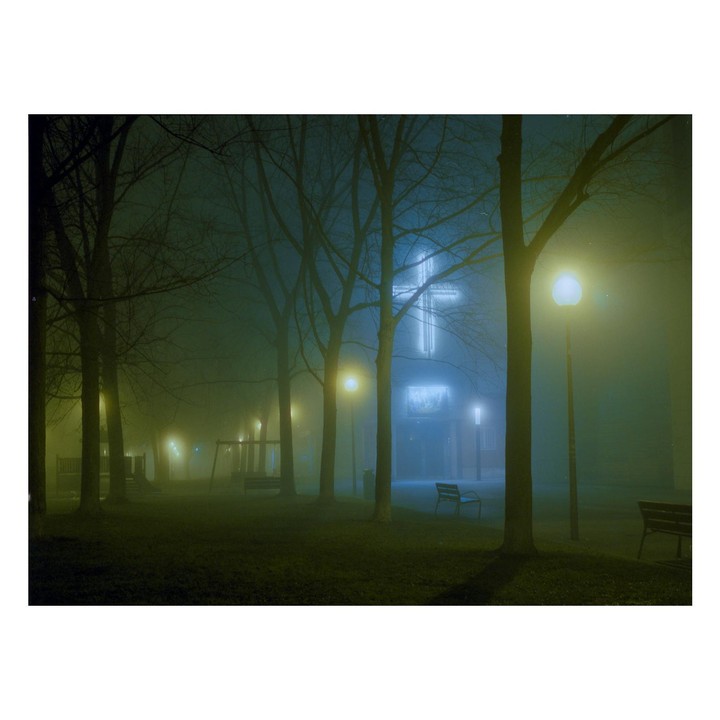 A park with bare trees and streetlights is shrouded in fog at night, with a glowing cross visible on a building in the background.