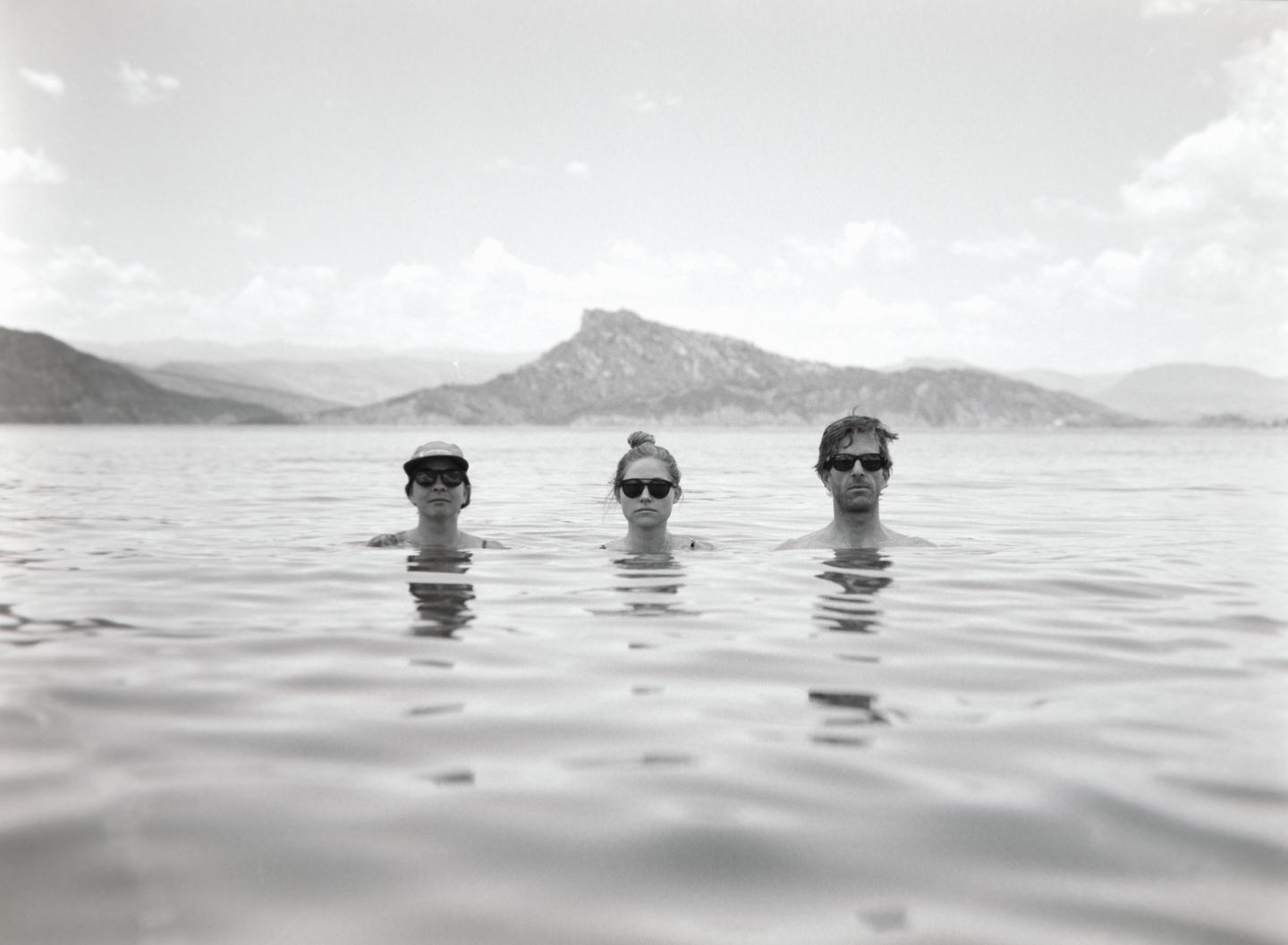 Three people wearing sunglasses are partially submerged in water with mountains in the background.