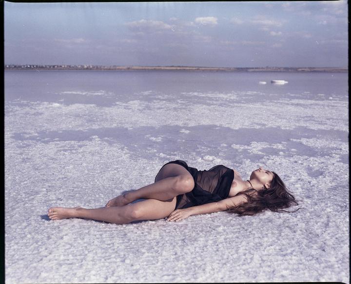 A woman lies on a salt flat with a body of water and a distant shore in the background.