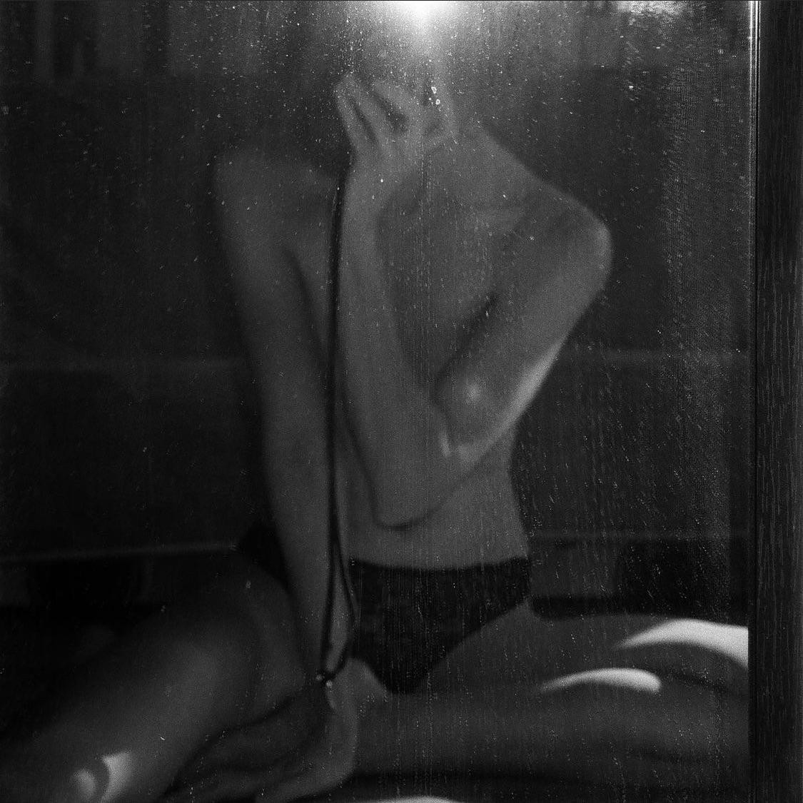A woman in underwear is reflected in a dusty mirror, with her arm covering her face.