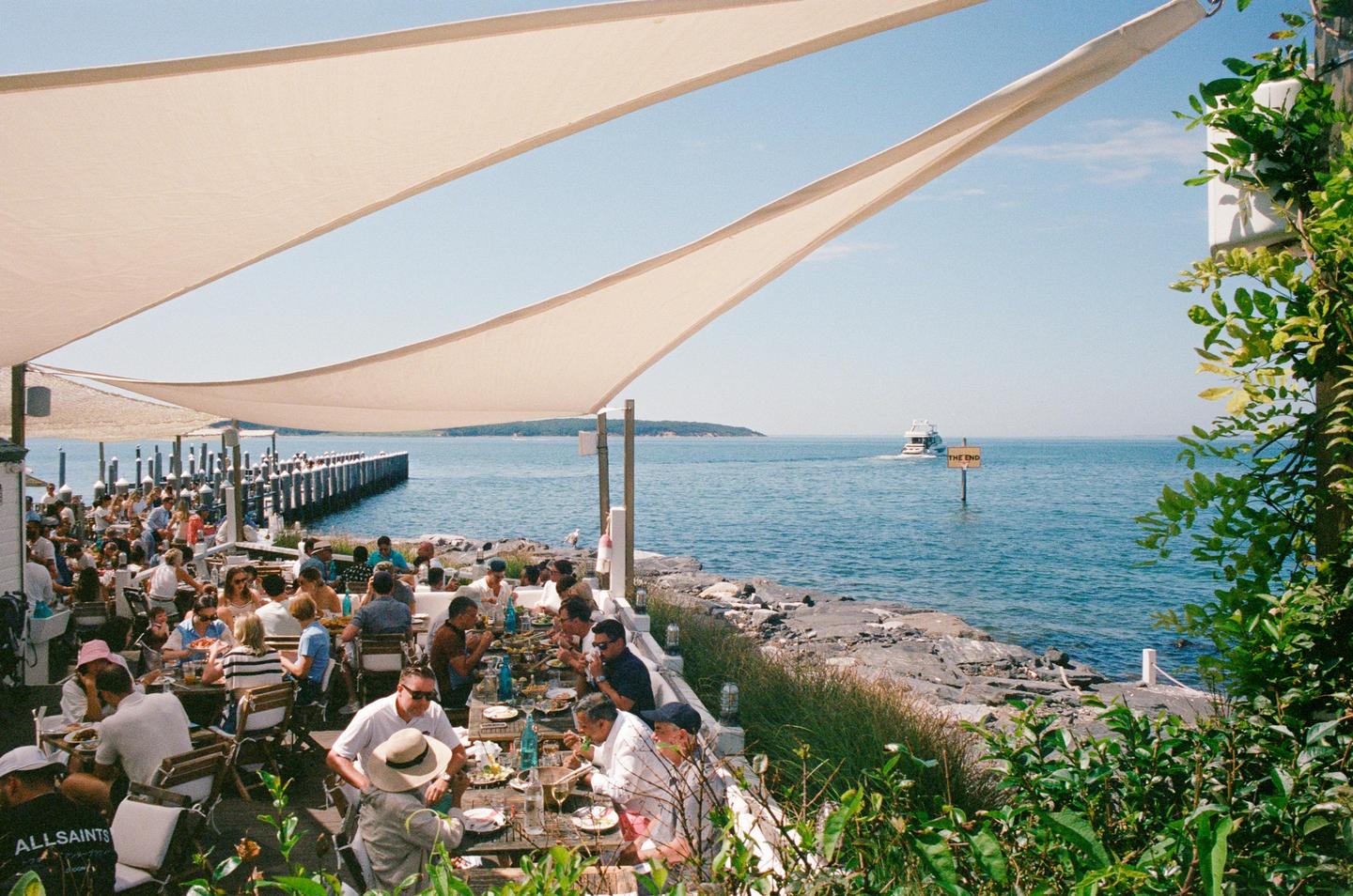 People eat lunch at tables under a large awning next to a body of water with a boat and a dock.