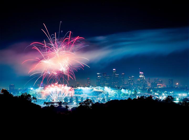 Fireworks explode over a city skyline at night, with silhouetted hills in the foreground.