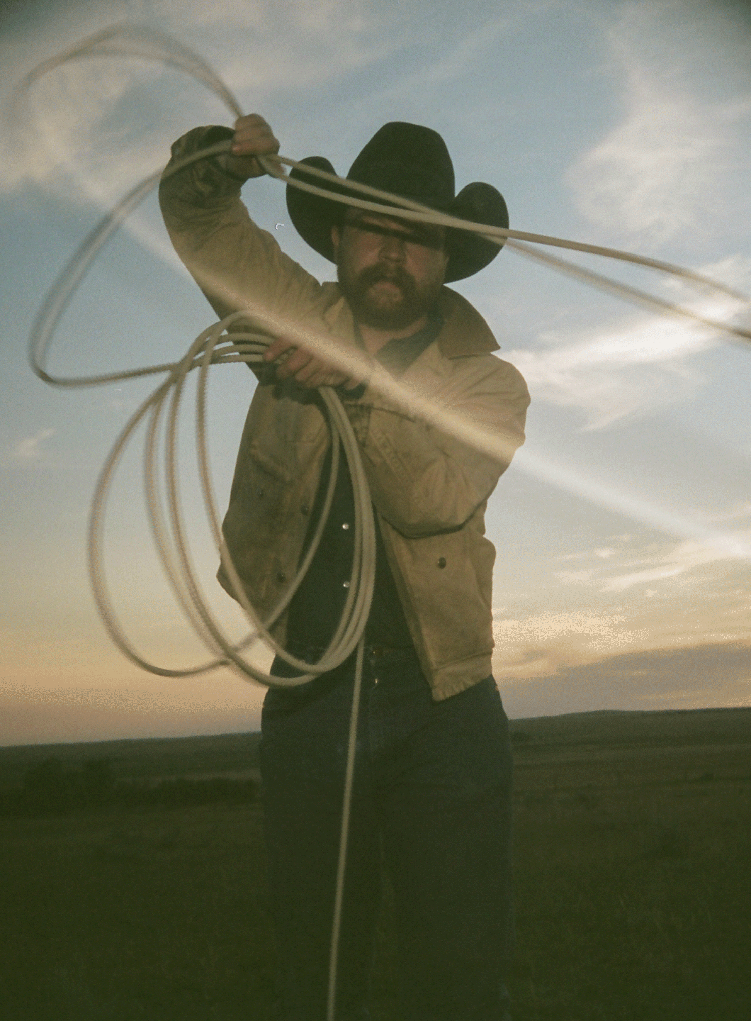 A man in a cowboy hat holds a lasso in a field at dusk.