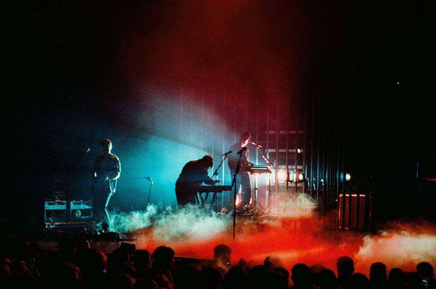 Three musicians perform on a stage illuminated by red and blue lights and smoke.