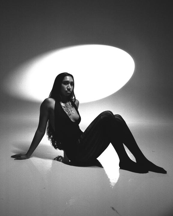 A woman in a dark top and striped pants sits on a reflective surface with a bright spotlight behind her.