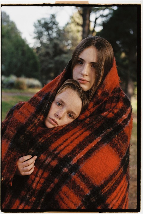 Two girls are wrapped in a red and black plaid blanket in a forest.