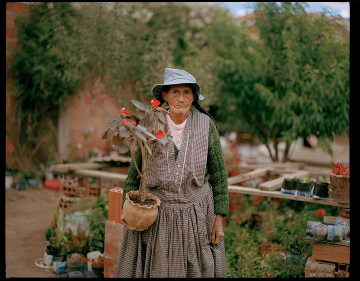 A woman in a hat and apron holds a potted plant with red flowers.