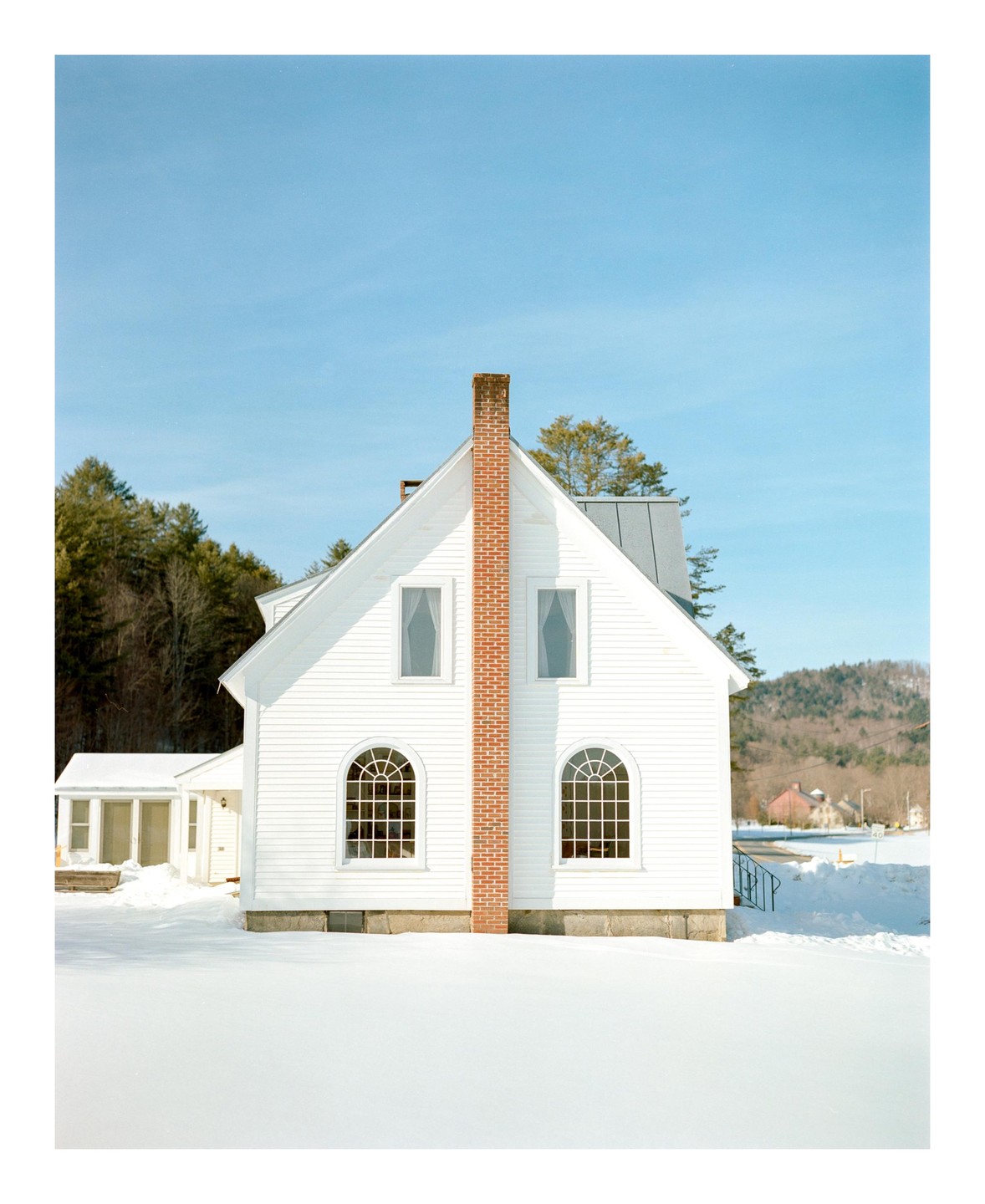 A white house with a prominent brick chimney stands in a snow-covered landscape under a clear blue sky.