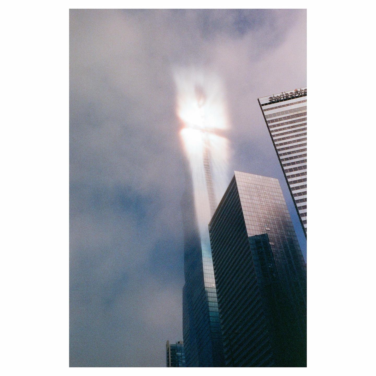 A tall skyscraper is partially obscured by clouds, with a bright light emanating from its upper section.