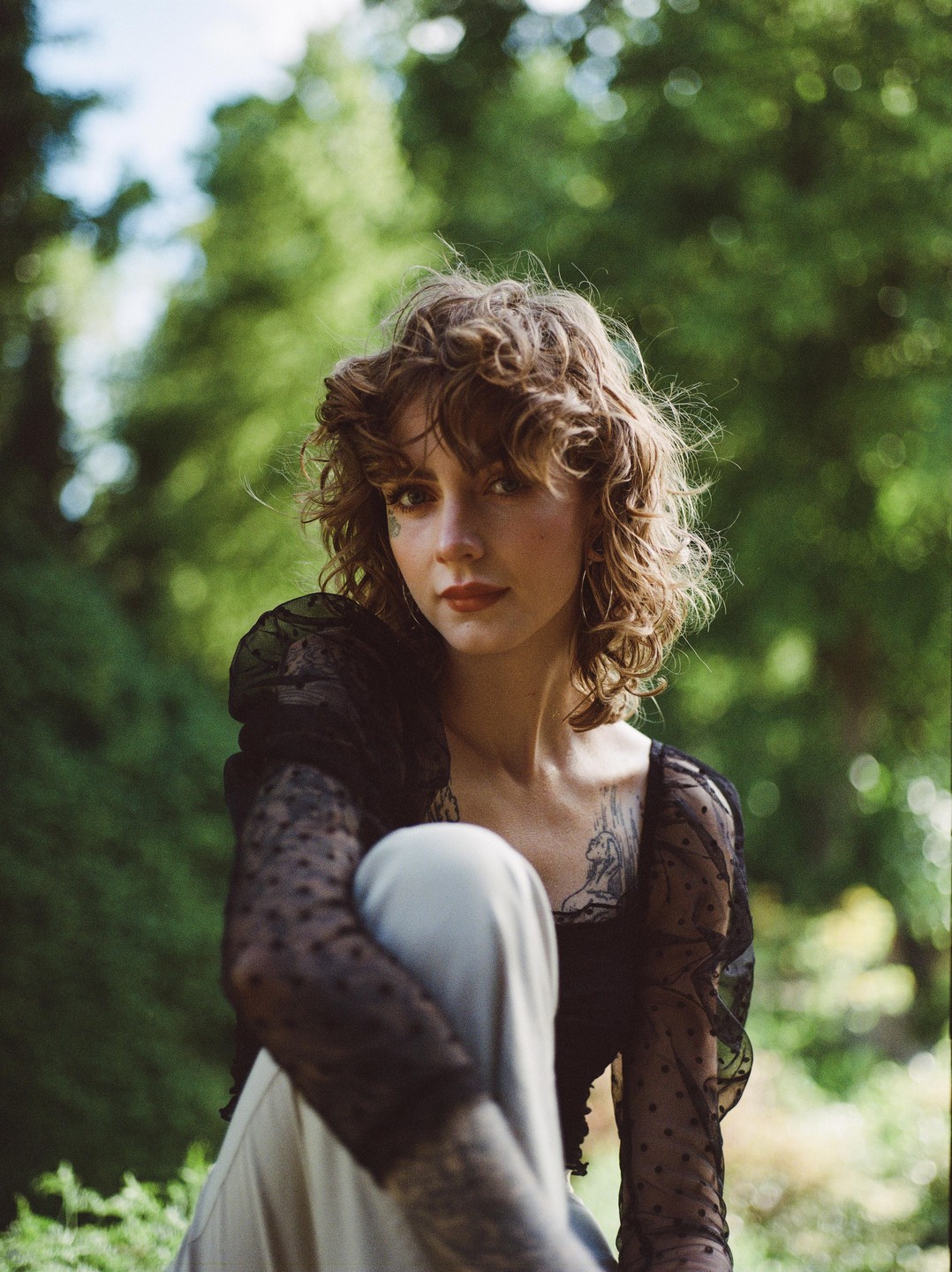A woman with curly hair and a black sheer top with polka dots sits outdoors.