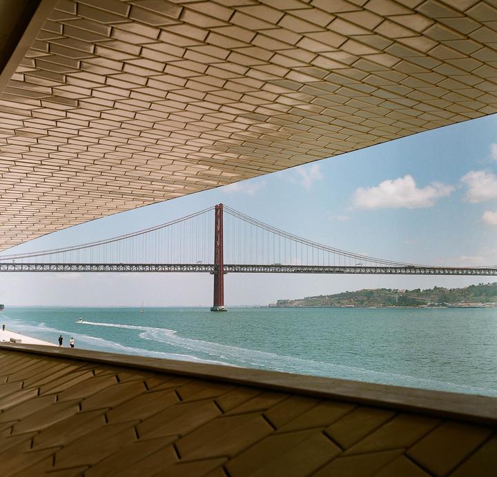 A red suspension bridge spans a wide river under a blue sky with scattered clouds, viewed from beneath a textured ceiling.
