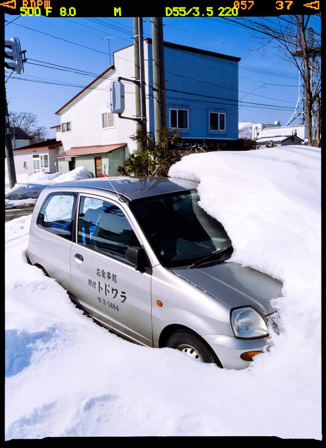 A silver car is partially buried in deep snow on a street with buildings and utility poles in the background.
