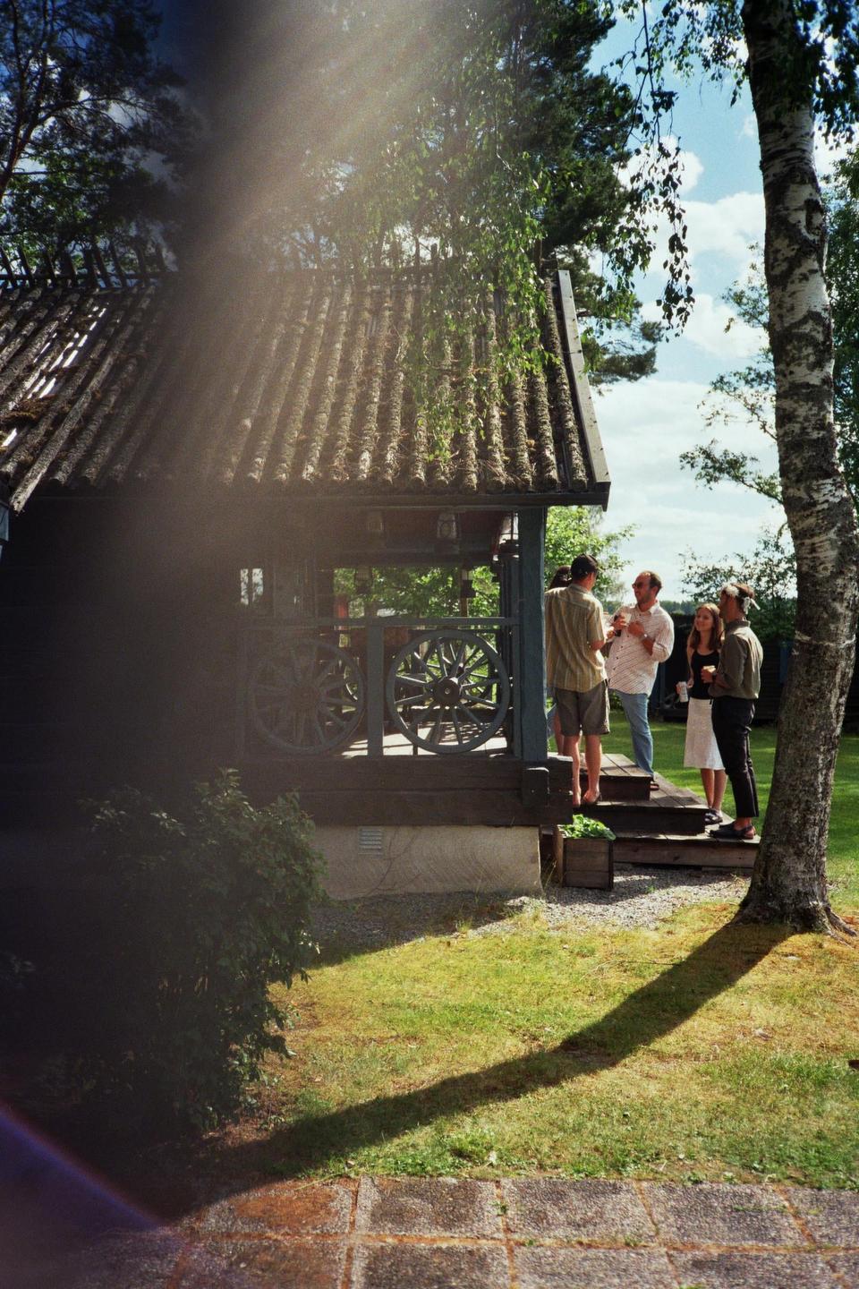 Four people stand on the porch of a cabin with a birch tree and grass in the foreground.