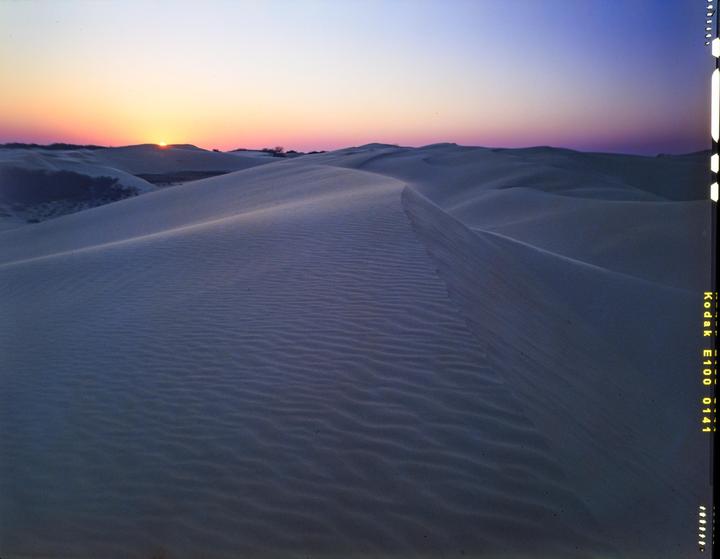 The sun sets over a vast expanse of sand dunes with ripples in the foreground.