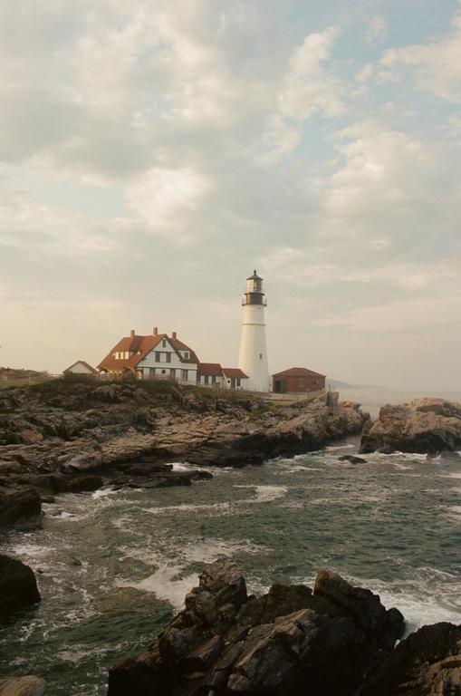 A white lighthouse stands on a rocky coastline next to a building with a red roof, with waves crashing against the shore.