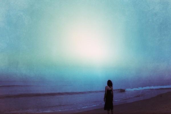 A woman stands on a beach looking out at the ocean under a bright, hazy sky.