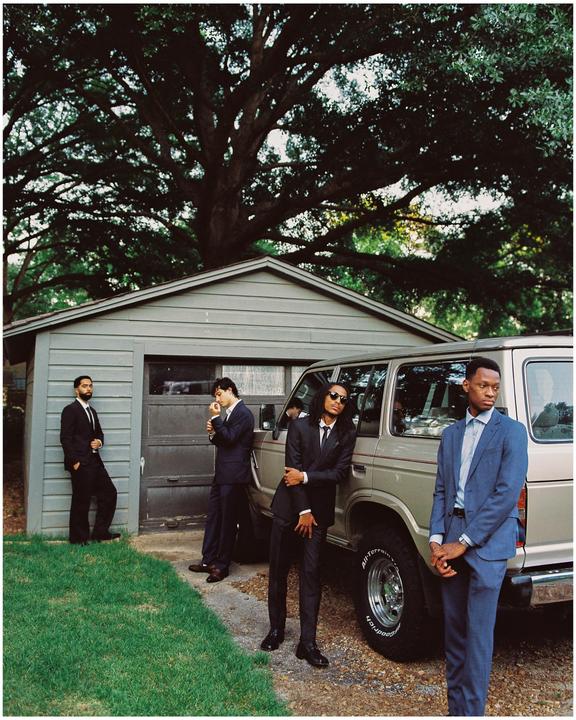 Four men in suits stand around a vehicle next to a garage with a large tree overhead.