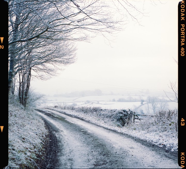 A snow-covered road leads through a winter landscape with bare trees and fields.